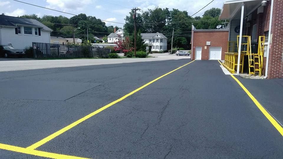 Black asphalt parking lot with yellow painted lines. Buildings and trees in the background.