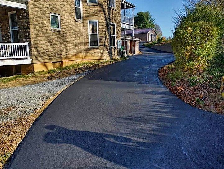 Asphalt driveway curves uphill next to a stone building. Sunlight casts a large shadow.