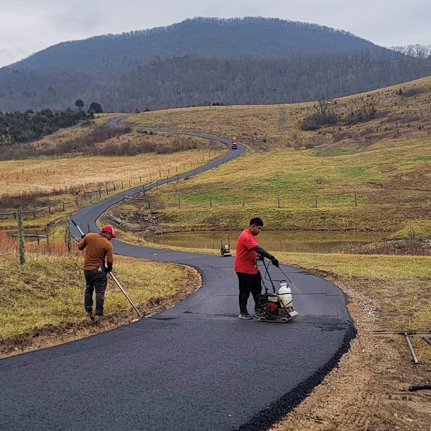 Two workers paving a winding road through a grassy field with a mountain backdrop.