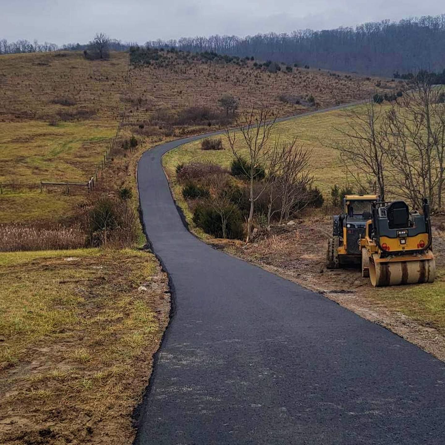 Paved road winding through a grassy field, construction equipment on the side, hills in the background.