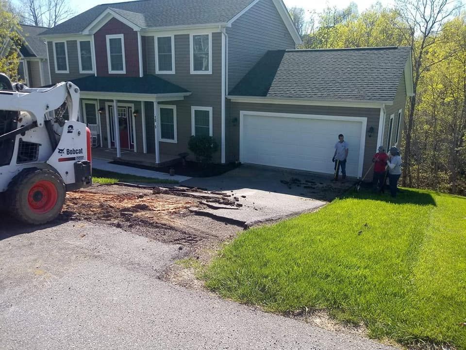 A Bobcat and workers near a house; driveway being worked on, green grass.