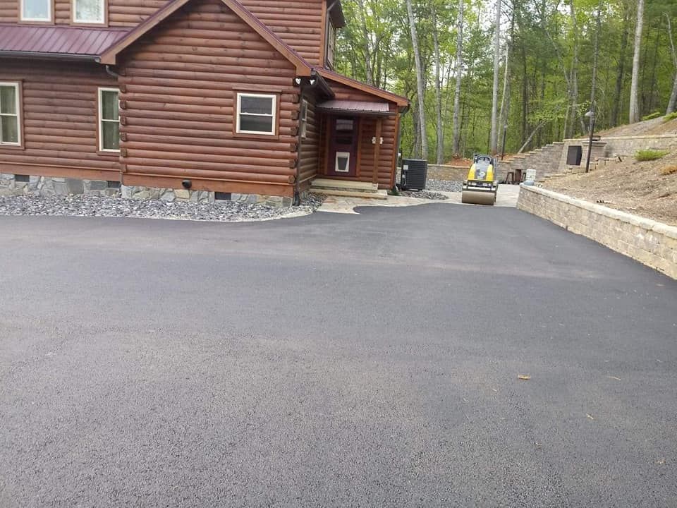 New asphalt driveway in front of a log cabin being smoothed by a roller; trees in the background.