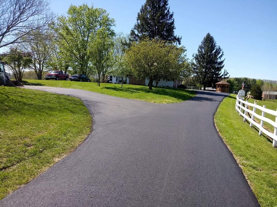 Paved driveway forks, leading to homes. Green grass surrounds with white fence on one side; blue sky.