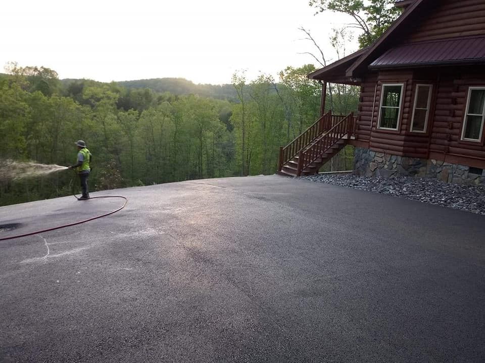 Man spraying sealant on asphalt driveway next to a log cabin with a mountain view.