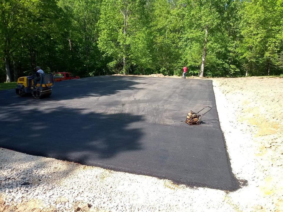 Asphalt paving of a parking area, with two pieces of machinery working; trees in the background.