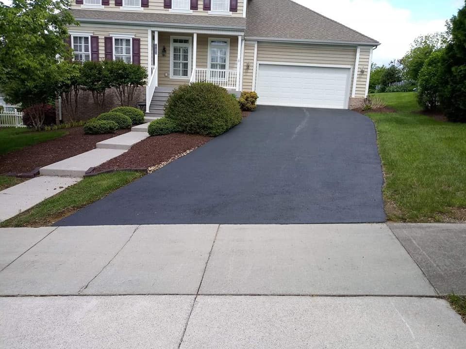 Driveway leading up to a two-story beige house with a garage; asphalt driveway is black, and sidewalk is concrete.