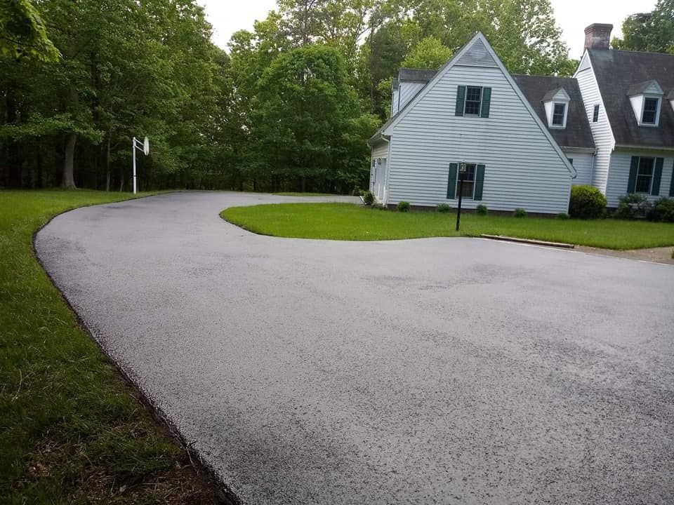 Asphalt driveway curves toward a white house with green shutters, trees in the background, and grassy lawn.