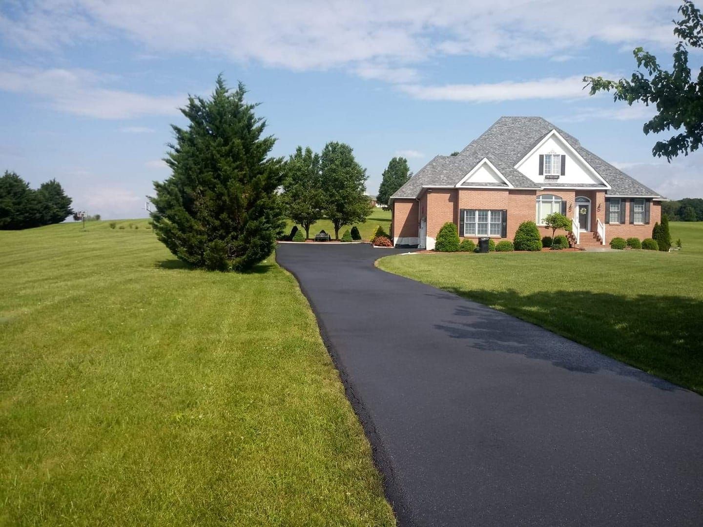 Asphalt driveway leads to brick house with a green lawn under a blue sky.