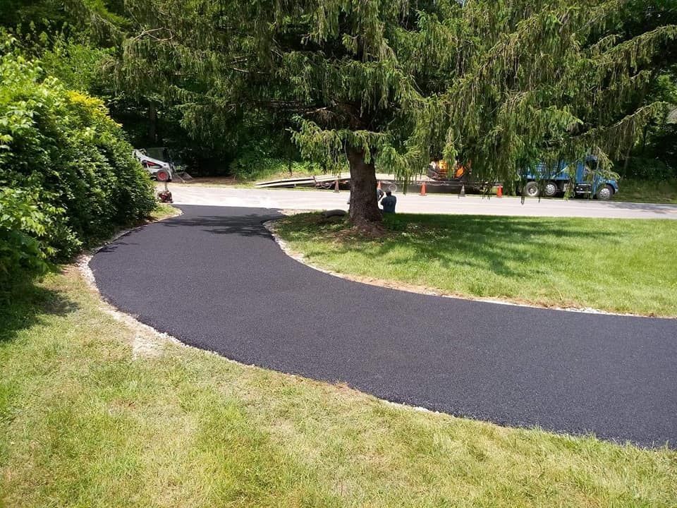 Newly paved black asphalt driveway curves through grassy lawn, tree in center.