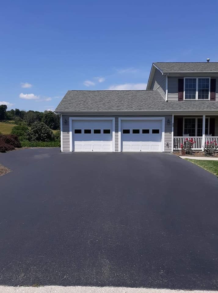 Asphalt driveway leads to a two-car garage with white doors, gray siding, and a house under a blue sky.