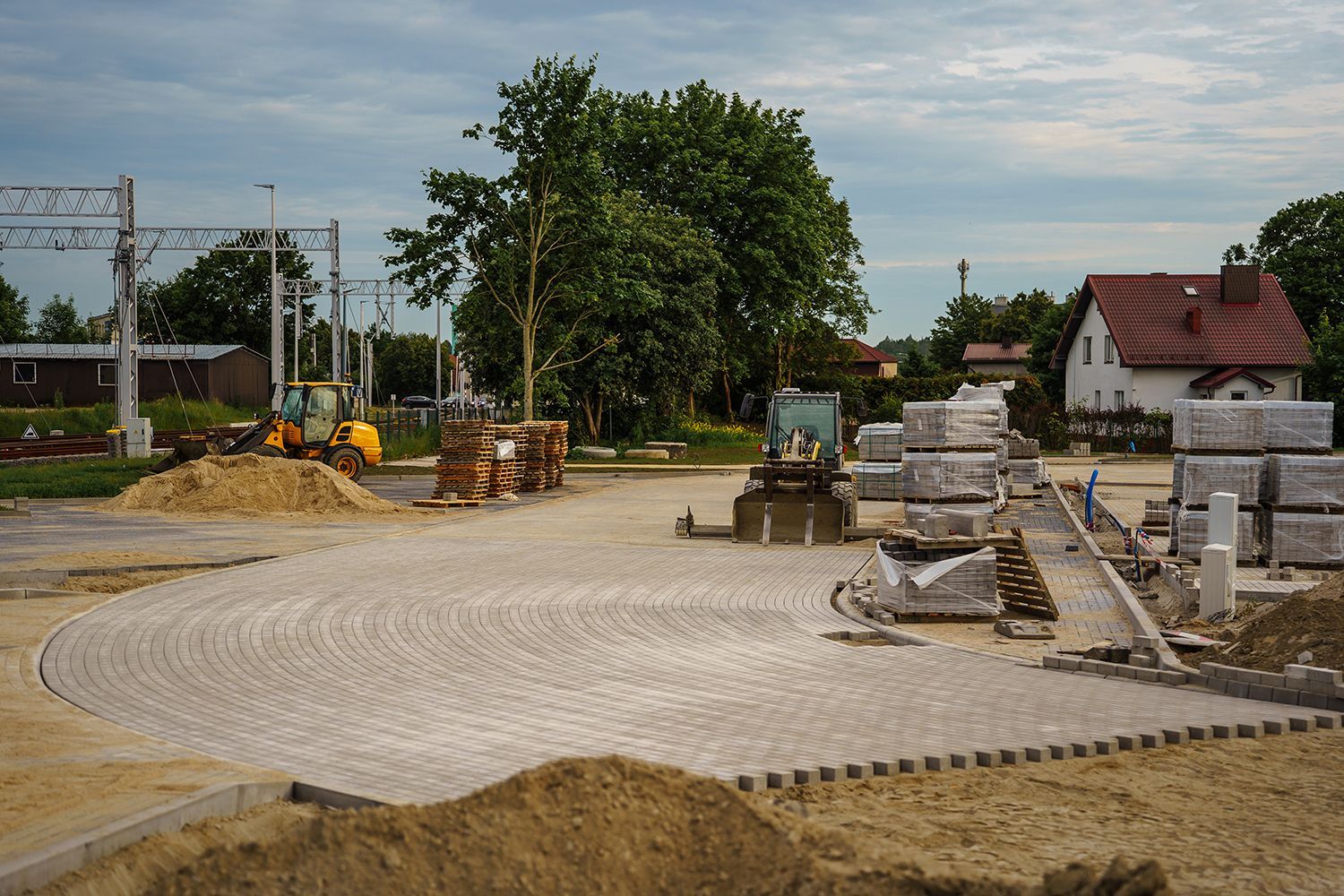 Road construction site with paved walkway, stacked materials, and trees near houses