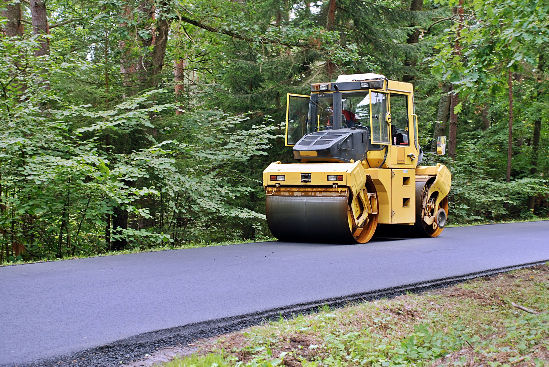 Yellow road roller compacting fresh asphalt on a paved roadway surrounded by trees. Yellow road roller compacting fresh asphalt on a paved roadway surrounded by trees.