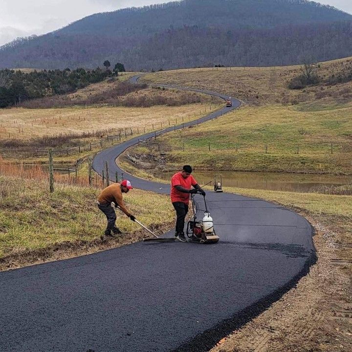 Two workers paving a winding asphalt road through a grassy landscape, near a pond and mountain.