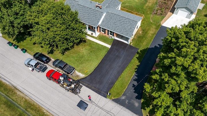 Aerial view: Asphalt driveway being paved; house with lawn, cars parked along street.