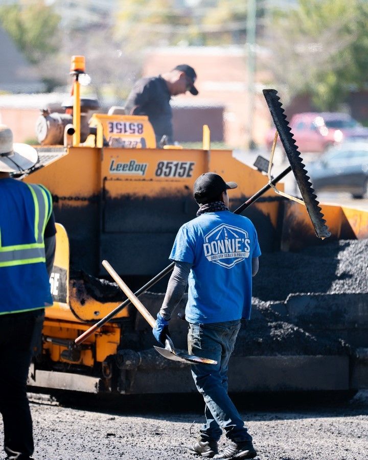 Road construction workers paving asphalt on a street. One worker carries tools, a machine in the background.
