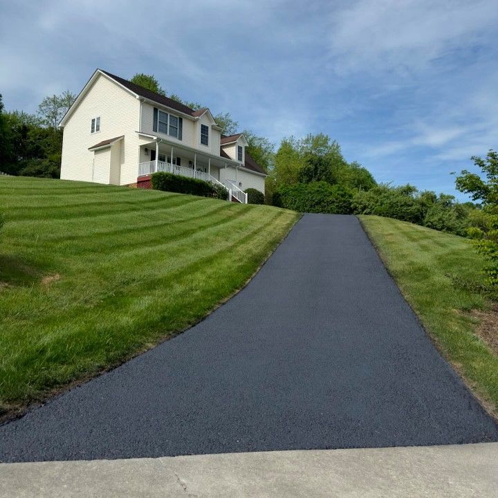 Newly paved asphalt driveway leading uphill to a two-story beige house on a grassy hill.