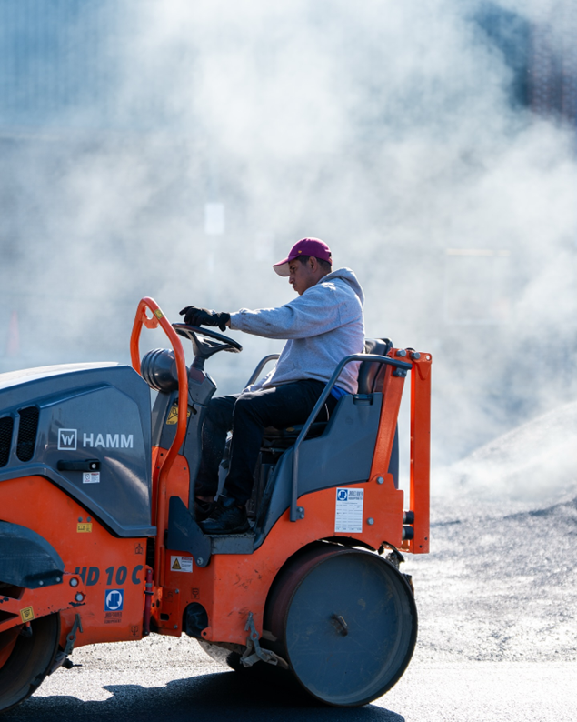 Man operating an orange HAMM HD 10 C roller on a road construction site.