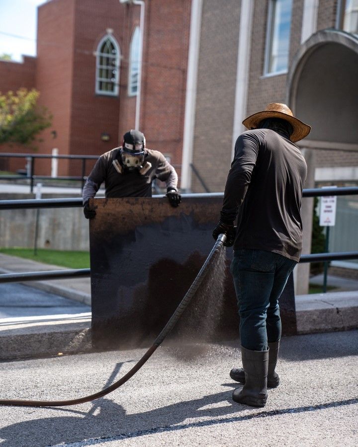 Two people cleaning a large, dark panel outdoors. One wears a respirator, the other a straw hat and boots, spraying water.