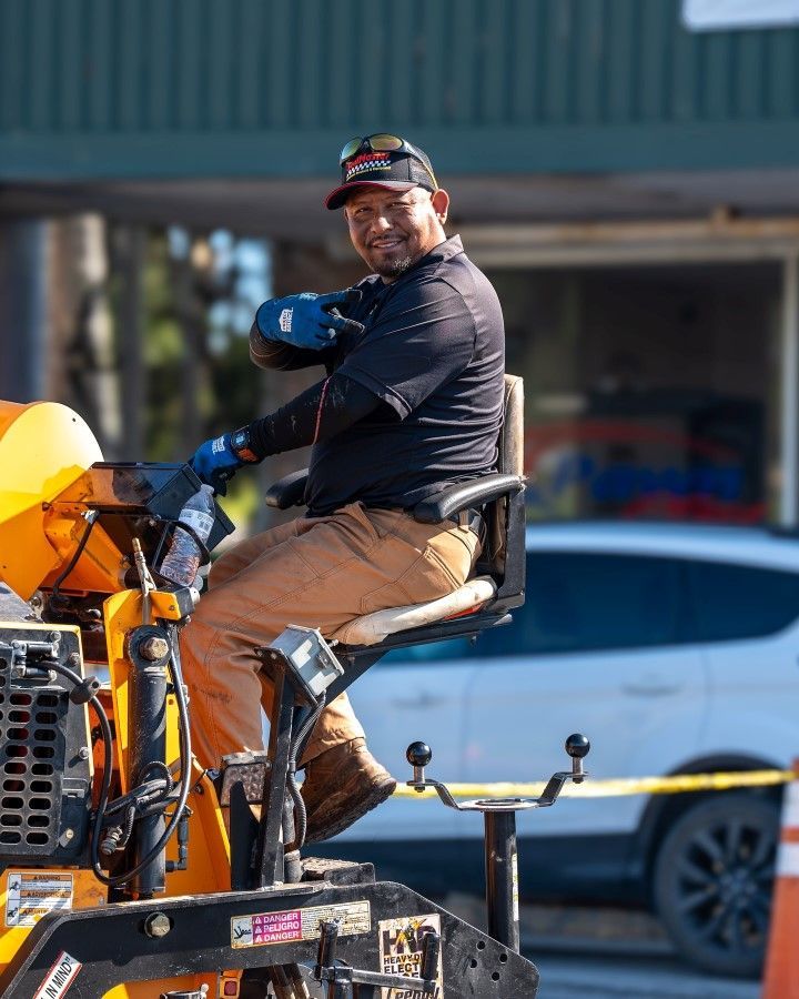 Man operating a yellow construction machine, smiling. Wearing a black shirt, brown pants, and a hat. Outdoors.