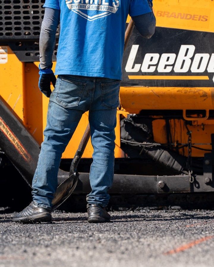 Construction worker stands next to paving equipment; wearing blue shirt, jeans, and gloves.