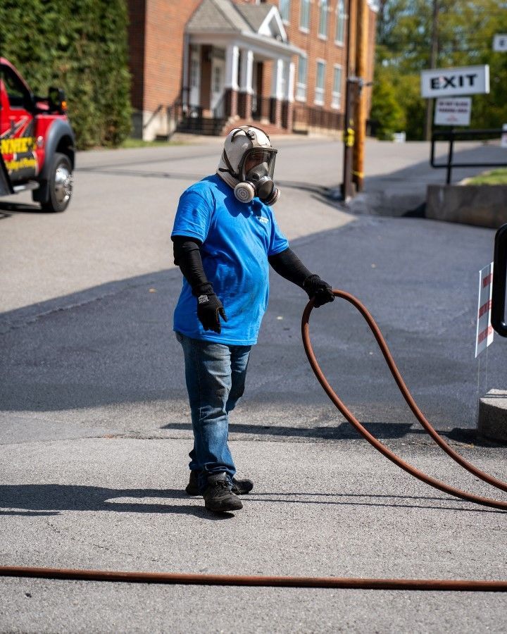 Person in a gas mask holds a hose on a paved street near a building and a tow truck.