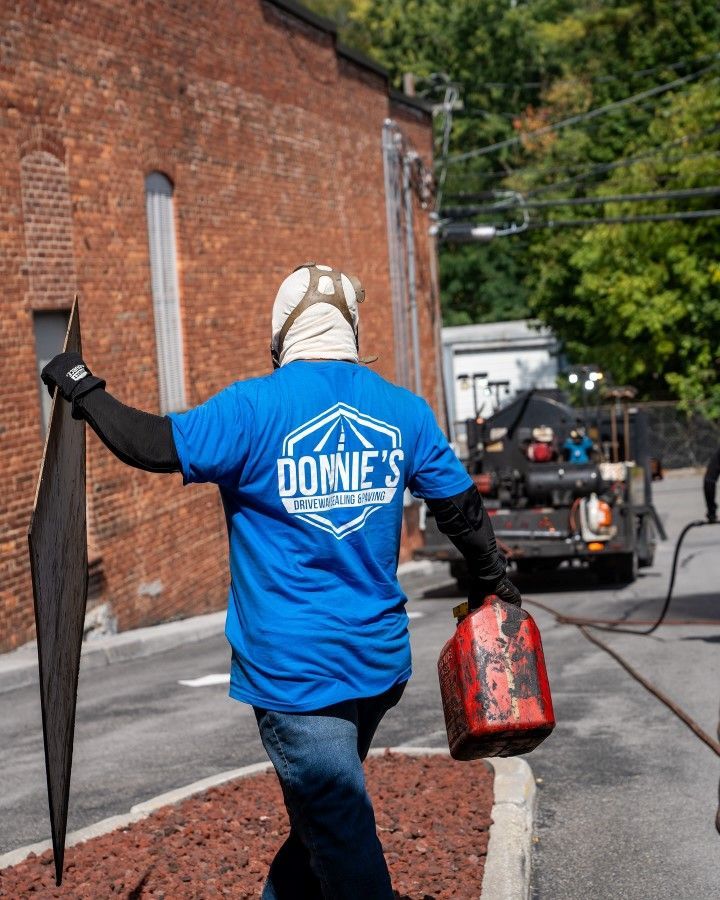 Person in blue shirt with a logo, holding a long tool and fuel canister, standing near a truck on a street.