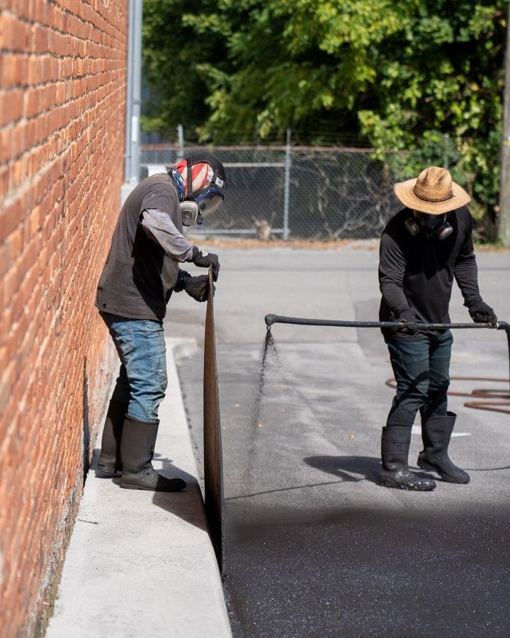 Two people spraying asphalt on a street next to a brick building.