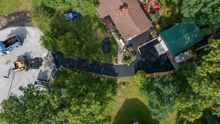 Aerial view of a house with a dark driveway, trees, and construction vehicles. Green roof on outbuilding.