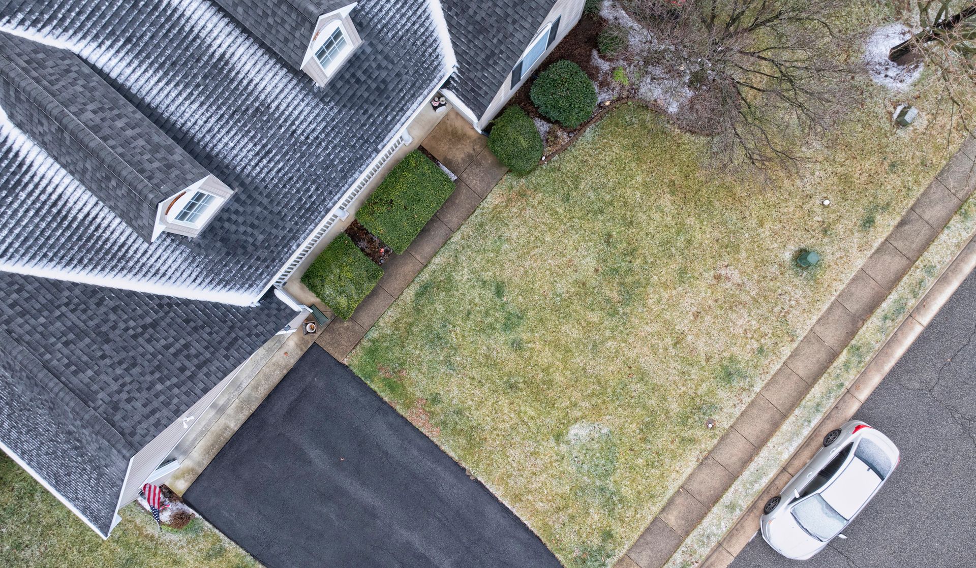 Overhead view of a house with a gray roof, small green bushes, a brown and green yard, and a car parked on the street.