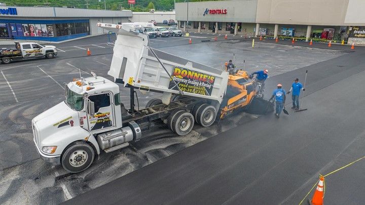 Asphalt paving: Dump truck unloading onto a paving machine with workers. Commercial lot in progress.