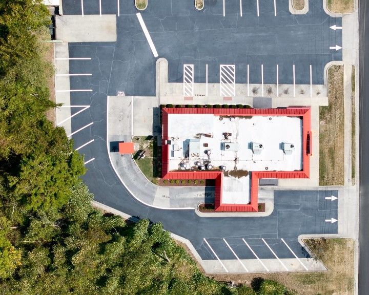 Aerial view of a restaurant with a red roof and parking lot. Black asphalt with white parking lines. Green trees on the side.
