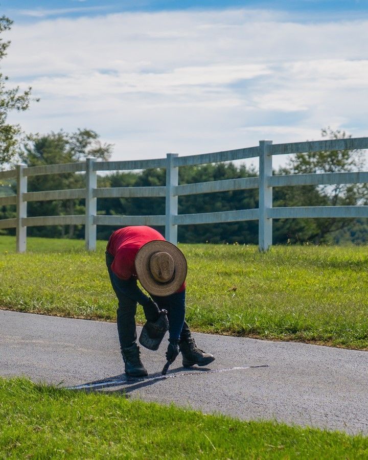 Person in red shirt and hat painting a line on a paved pathway, white fence and green grass in background.