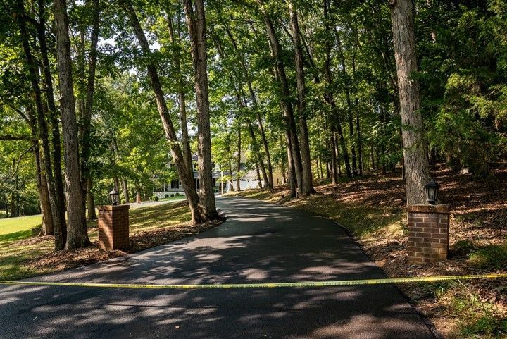 Paved driveway curves into a wooded area, bordered by trees and brick pillars. Yellow caution tape across the foreground.