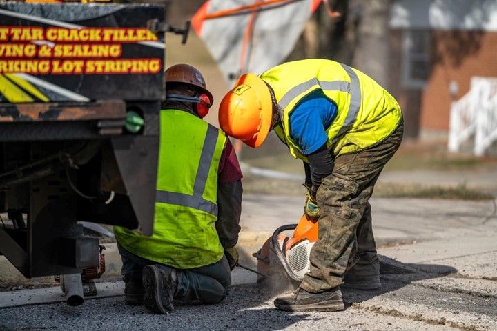 Two workers in safety vests and helmets, one kneeling and sawing pavement near a truck.