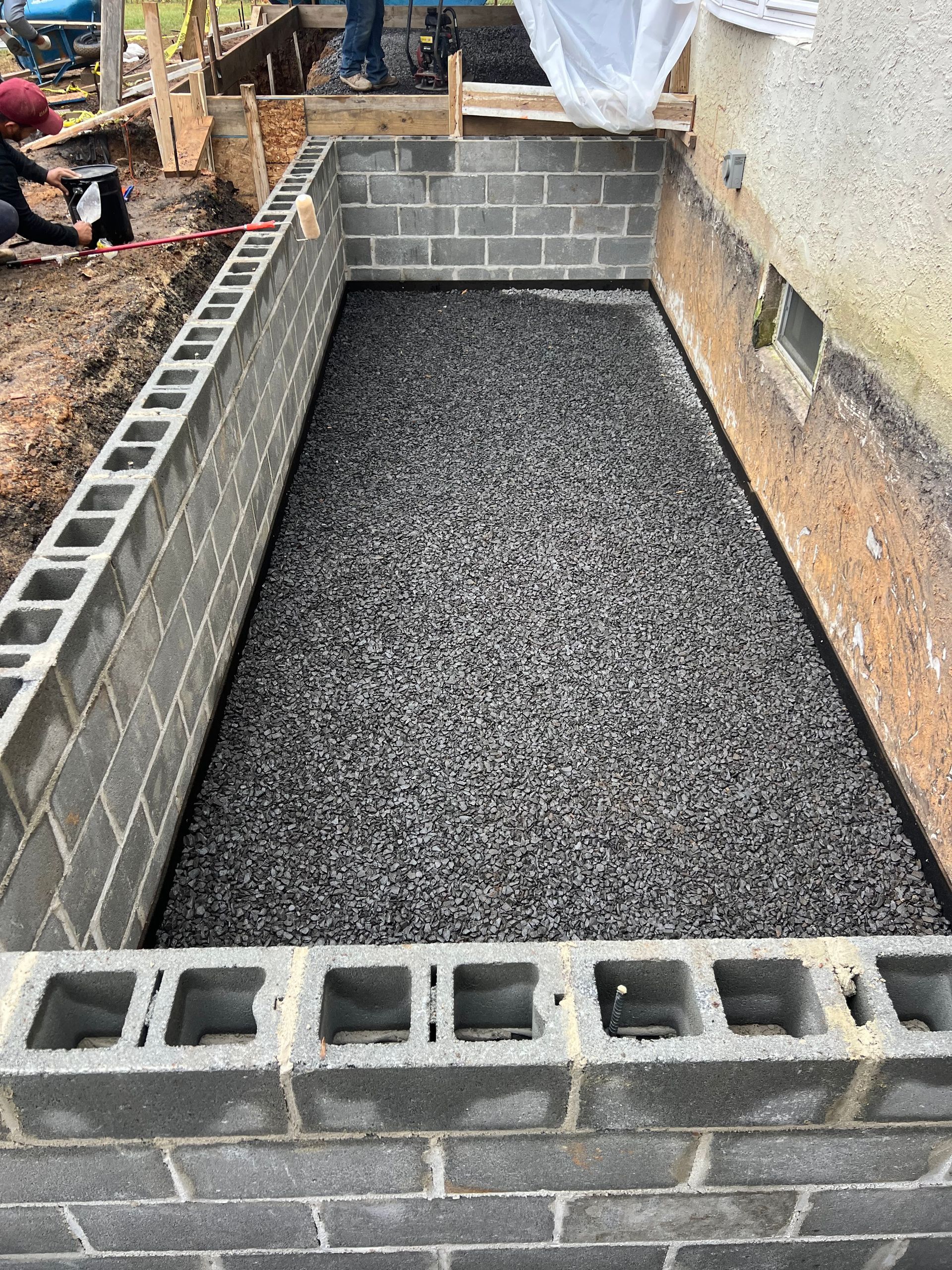 Rectangular cinder block foundation with gravel base, next to a wall. Construction scene.