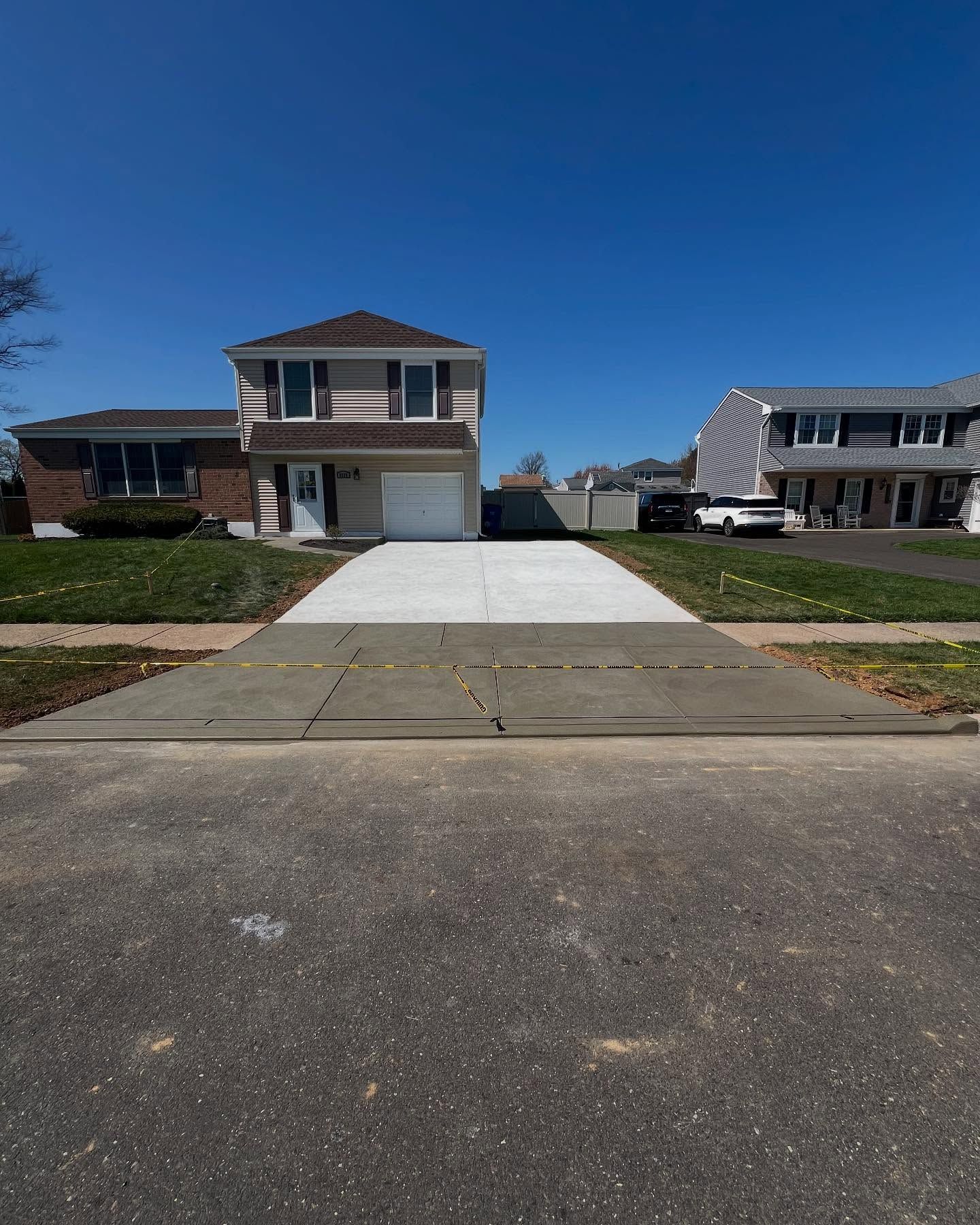 Two-story beige house with a concrete driveway. A street with a visible road in the foreground. Blue sky.