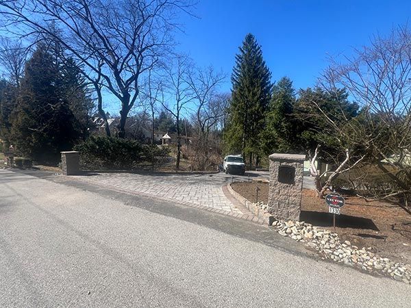 Gravel driveway entrance with stone pillars, a car, and trees under a clear, blue sky.