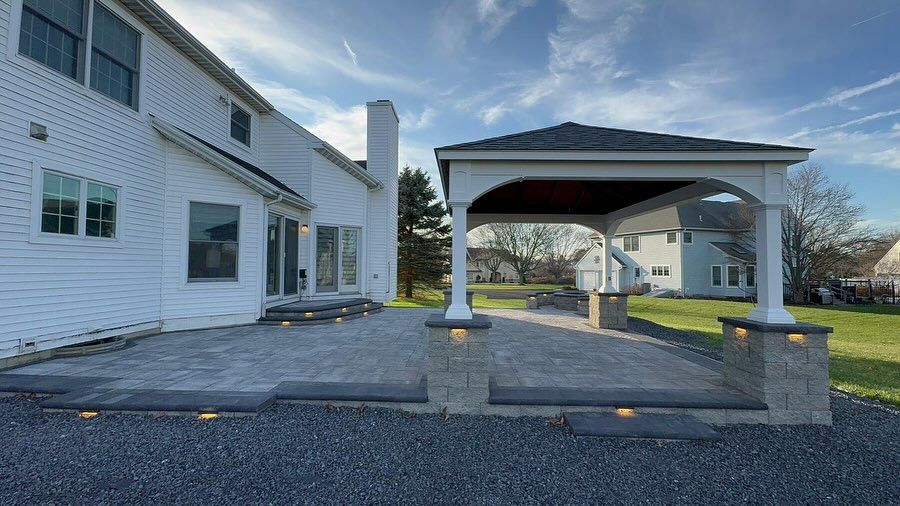 White house with paved patio, connected to a covered pavilion with pillars. Blue sky.