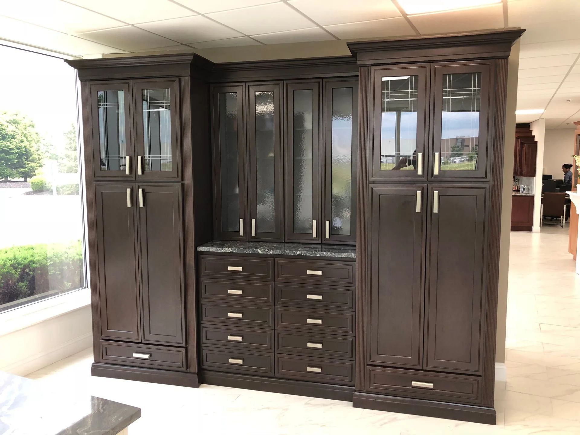 Dark wood display cabinet with glass doors and drawers in a showroom or lobby.