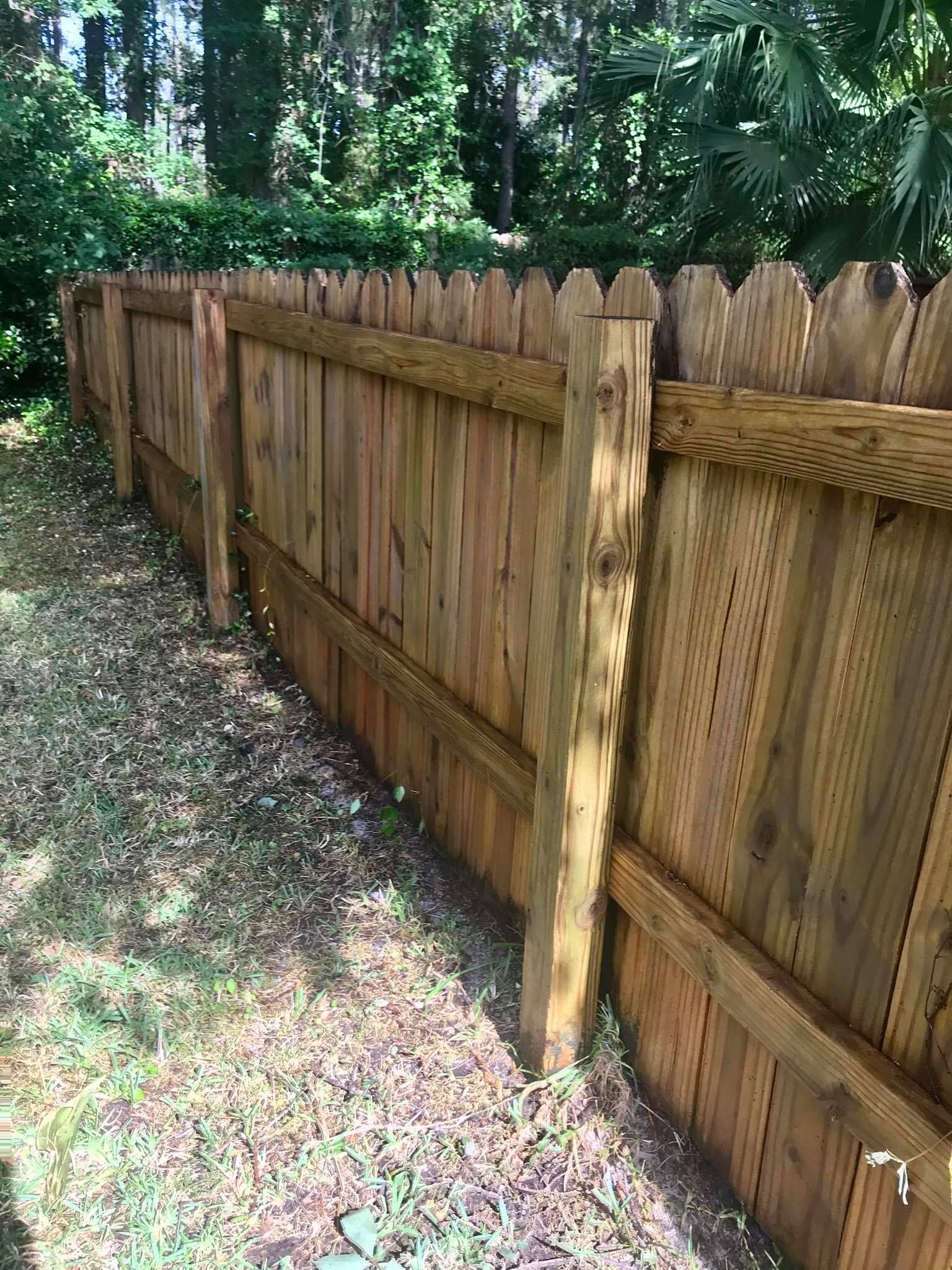 Wooden fence in a yard, angled view. Brown wood, pointed tops, green grass, and trees in the background.
