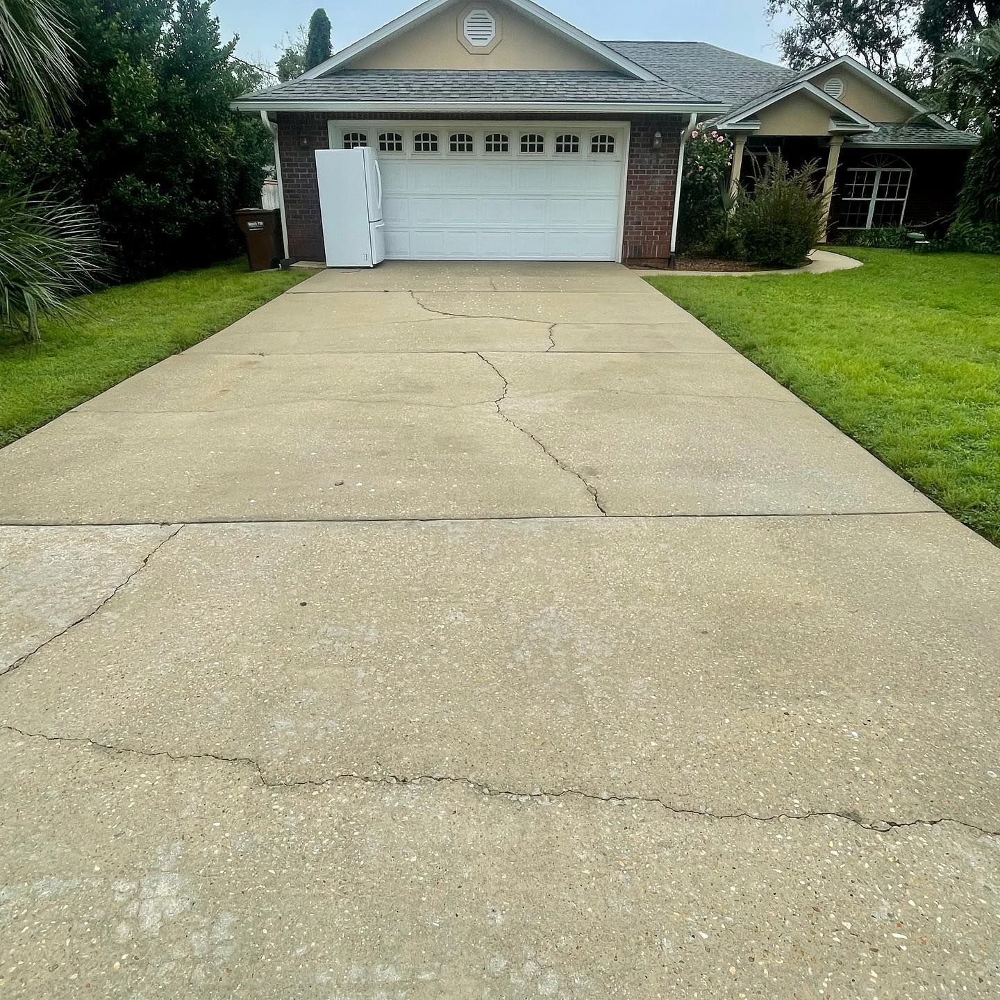 Cracked concrete driveway leading to a house with a white garage door, surrounded by grass and trees.