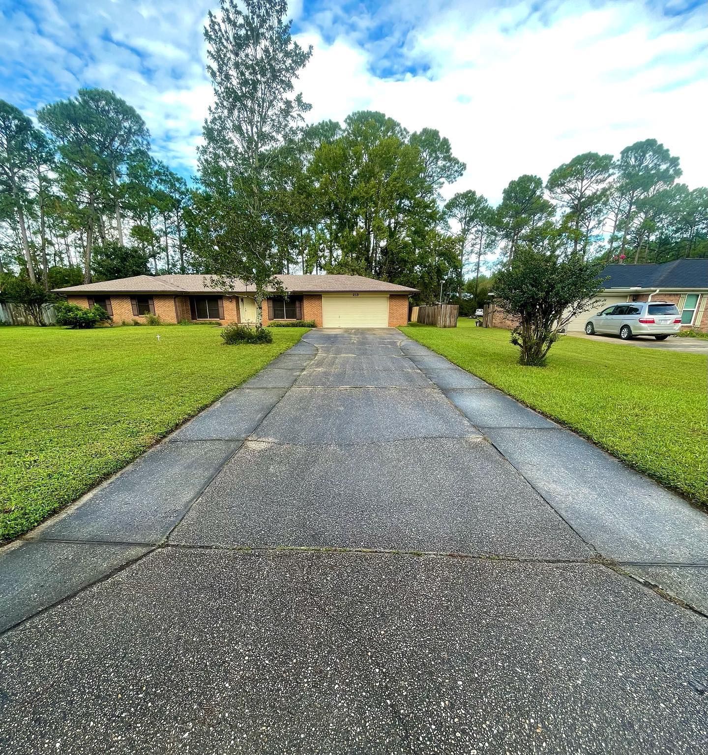 Brick ranch house with long concrete driveway, green lawn, and trees under a cloudy sky.