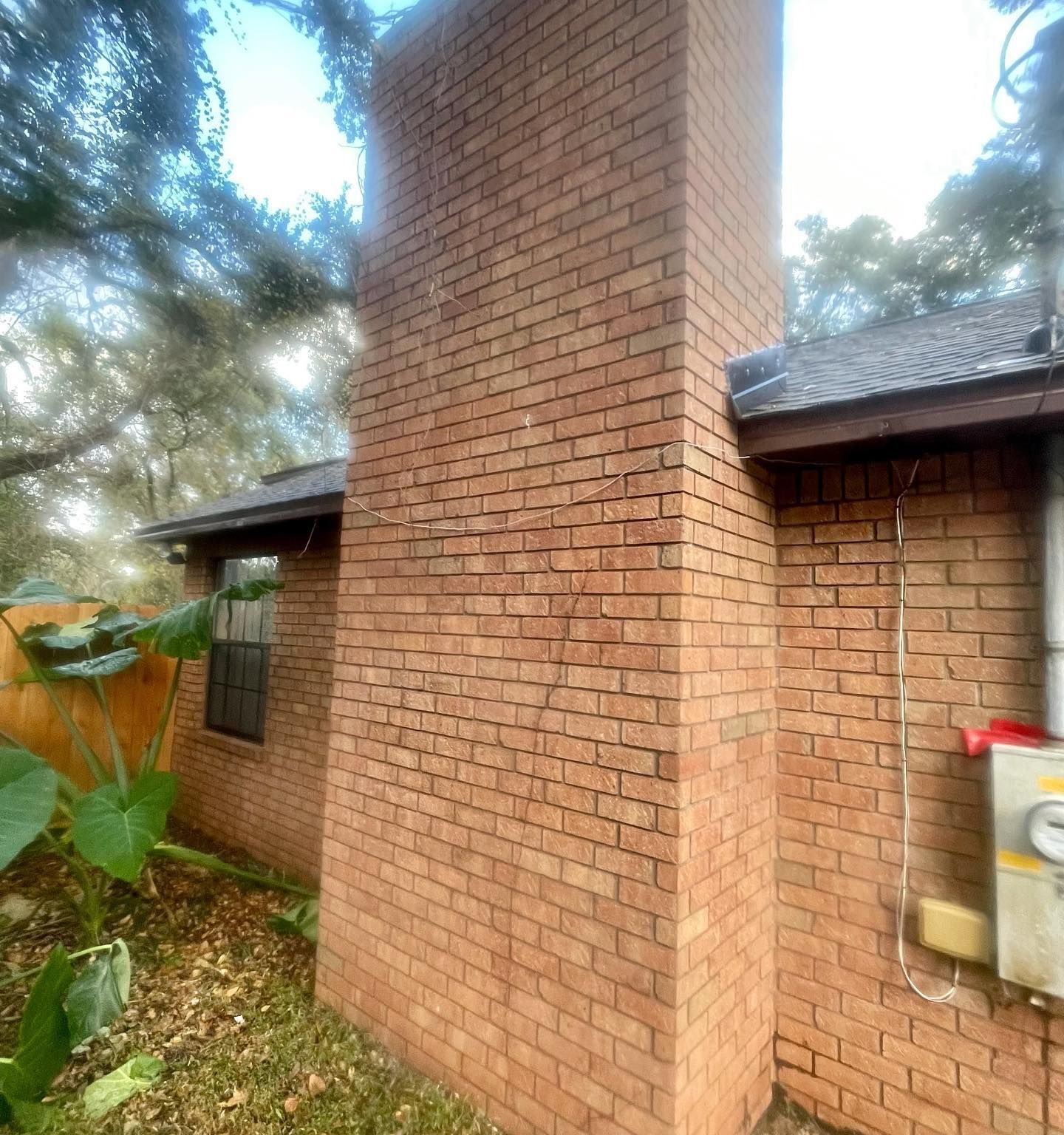 Brick chimney on a house exterior; tan bricks, brown roof, and electrical box on the wall.
