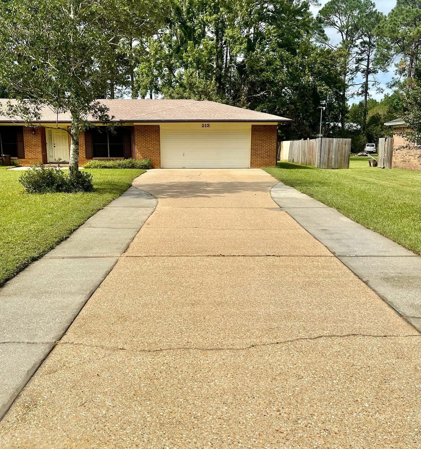 A one-story brick house with a concrete driveway and sidewalk, grass on either side, and trees in the background.