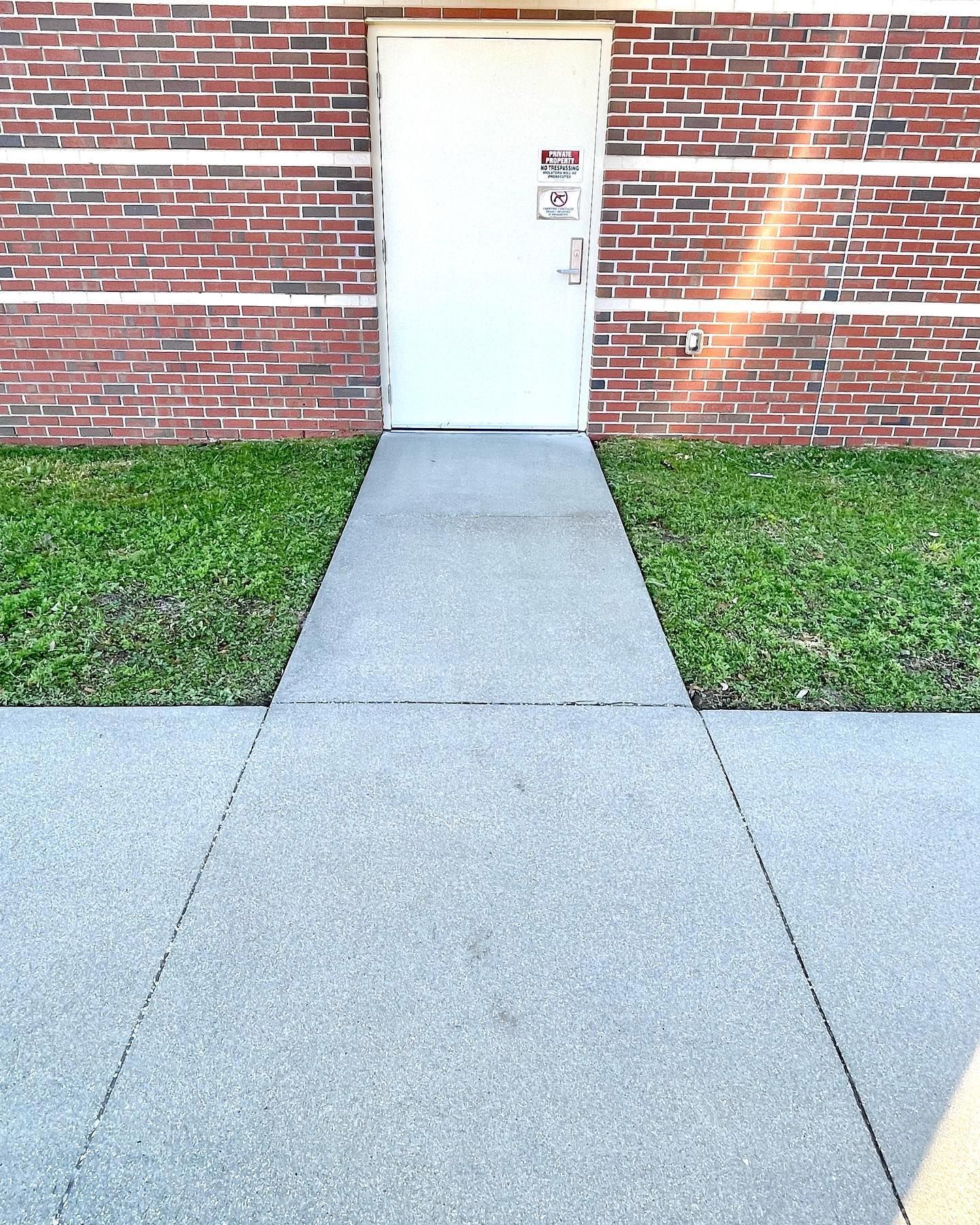Concrete walkway leading to a white door in a brick building. Grass borders the walkway on either side.