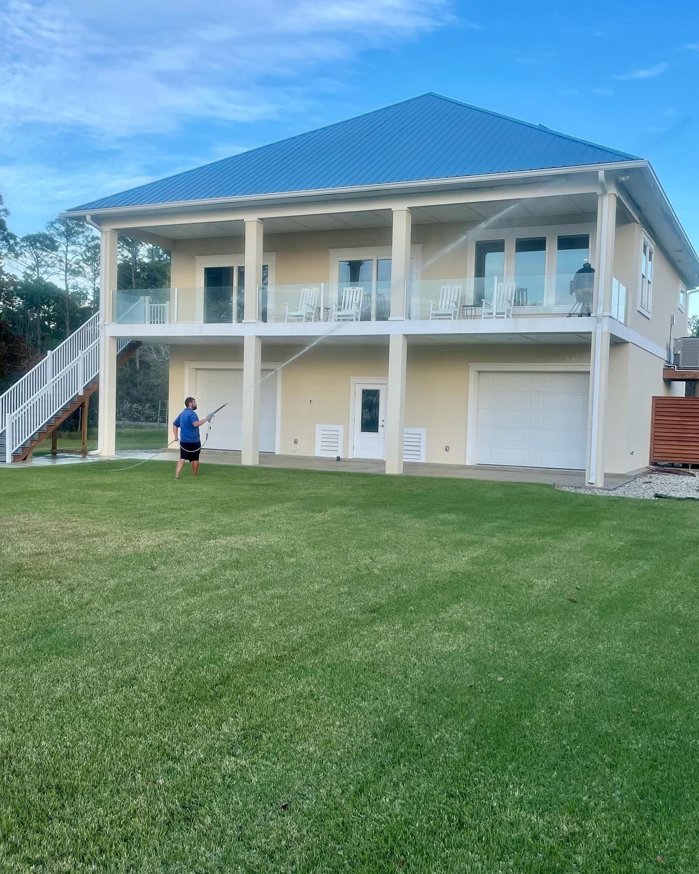 Two-story beige house with blue roof and balconies overlooking a green lawn. A person is outside holding a stick.
