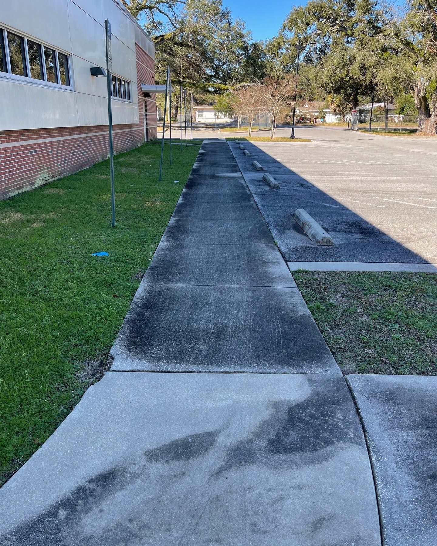Concrete sidewalk next to a building and parking lot, with patches of grass and blue sky.
