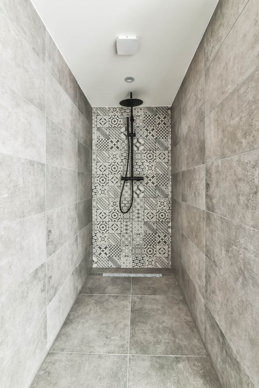 Narrow tiled walk-in shower with rainfall showerhead and patterned back wall.