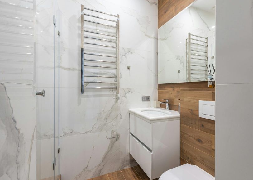A modern bathroom with white marble-like tiles, a white vanity, a toilet, and a silver heated towel rack.