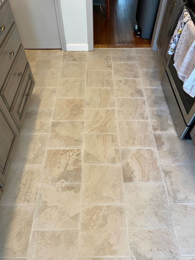 Beige tiled kitchen floor with cabinets and a glimpse into another room with hardwood flooring.